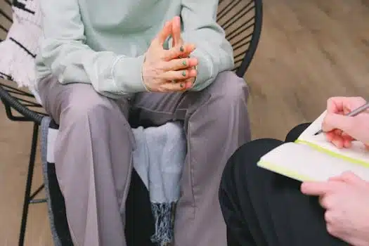 A person sitting in a chair with clasped hands, wearing a light-coloured sweatshirt and pants, while another person takes notes in a notebook, suggesting a counselling or therapy session.