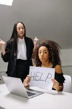 A woman holding a 'HELP' sign while sitting at a desk with a laptop, and another person standing beside her in an office setting.