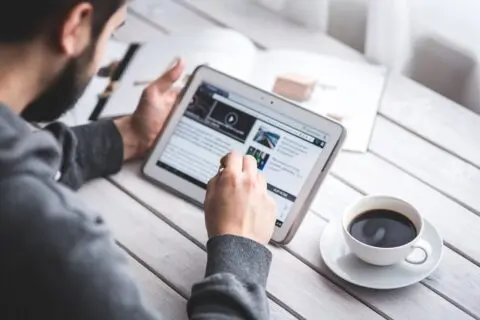 A person using a tablet while sitting at a wooden table with a cup of coffee, browsing a website or reading an article in a bright, modern setting.